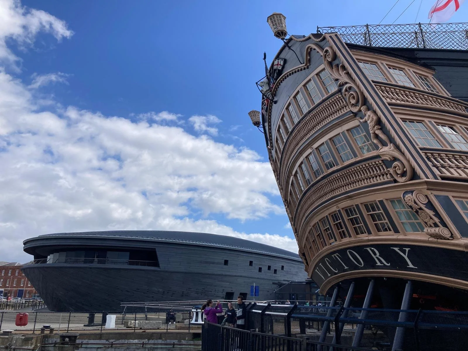 A large old-fashioned wooden ship named Victory with ornate details and a modern building with a curved roof in the background. There are a few people in the foreground near a fence.