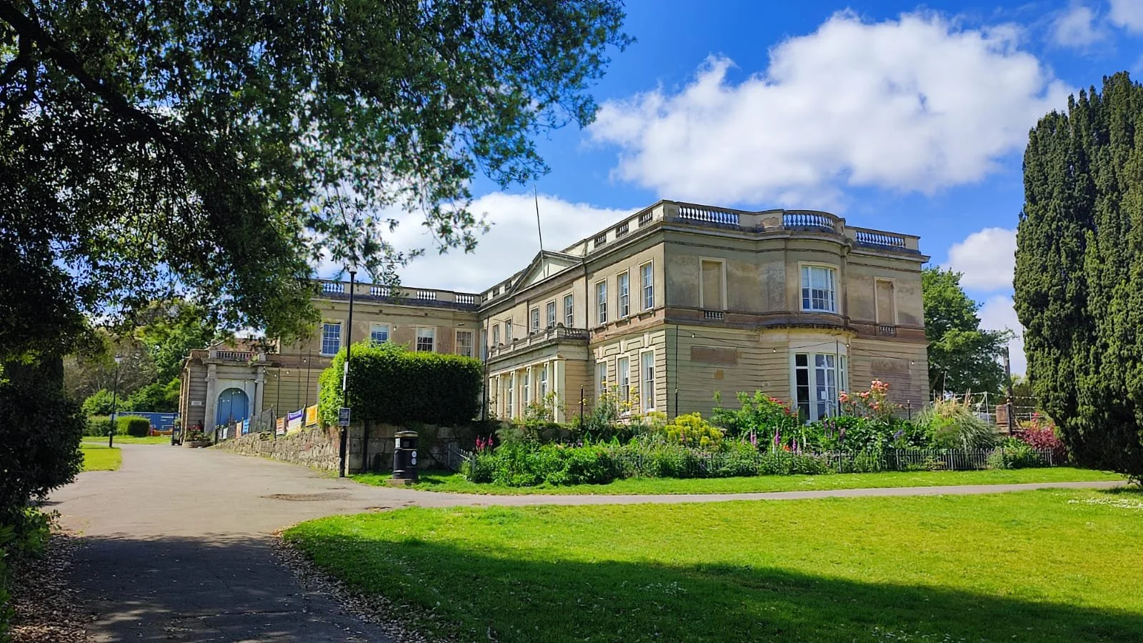 Large, historic mansion with beige stone exterior, multiple windows, and a garden with flowers in front, set in a lush green park with a pathway and trees, under a partly cloudy sky.