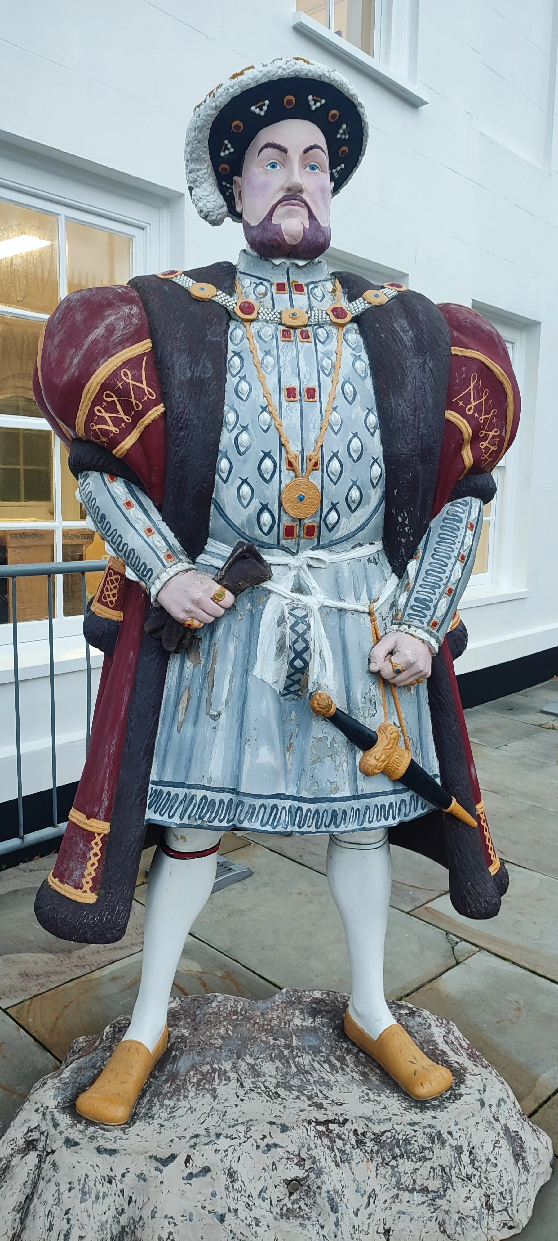 A painted wooden statue of a man dressed as a royal or noble figure from historical times, wearing a hat, ornate clothing, and holding a sword. The statue stands outdoors in front of a white building.