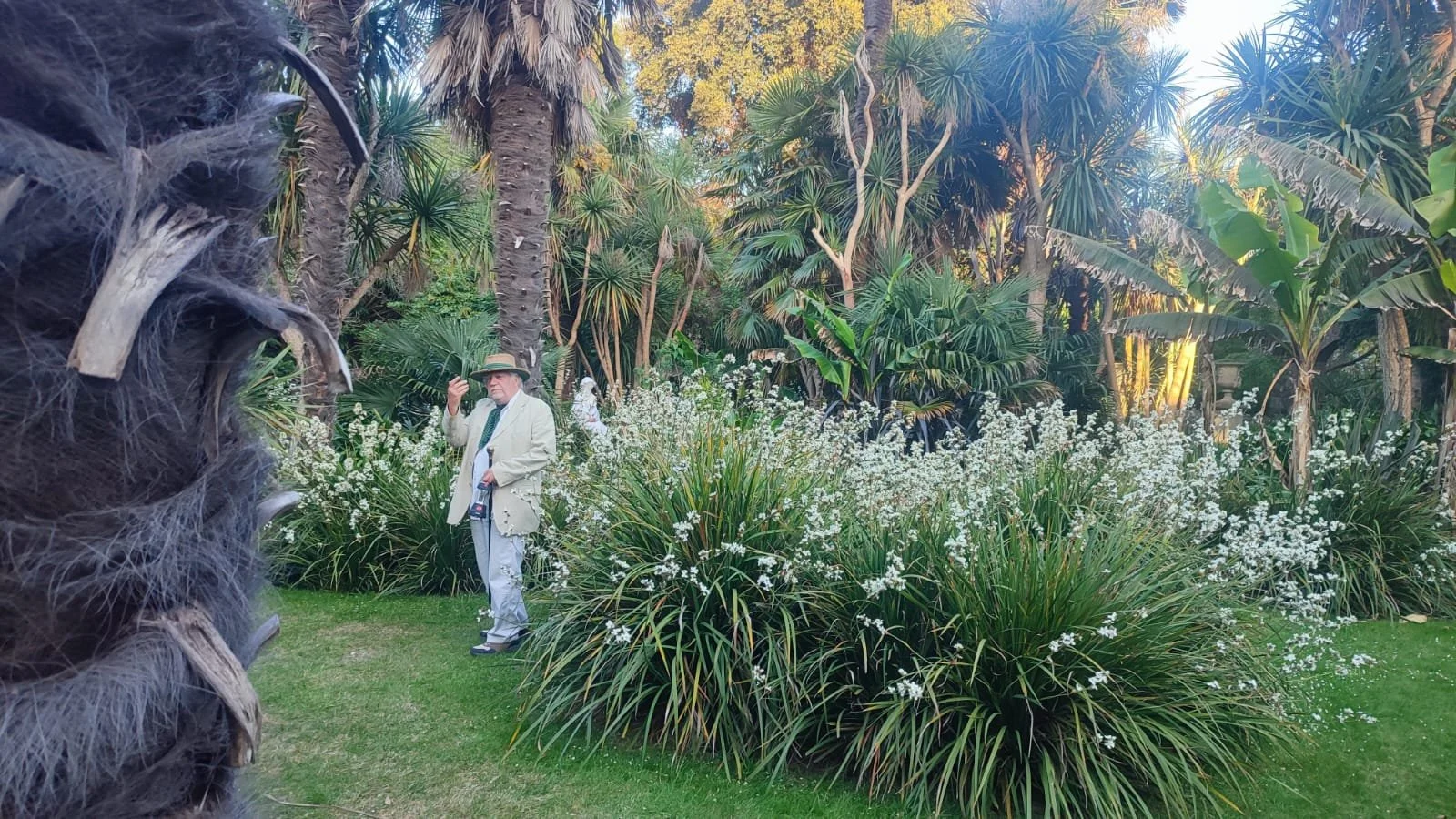 A man in a light-colored coat and hat standing in a lush garden with tall palms and blooming white flowers, holding a camera and a cigarette.