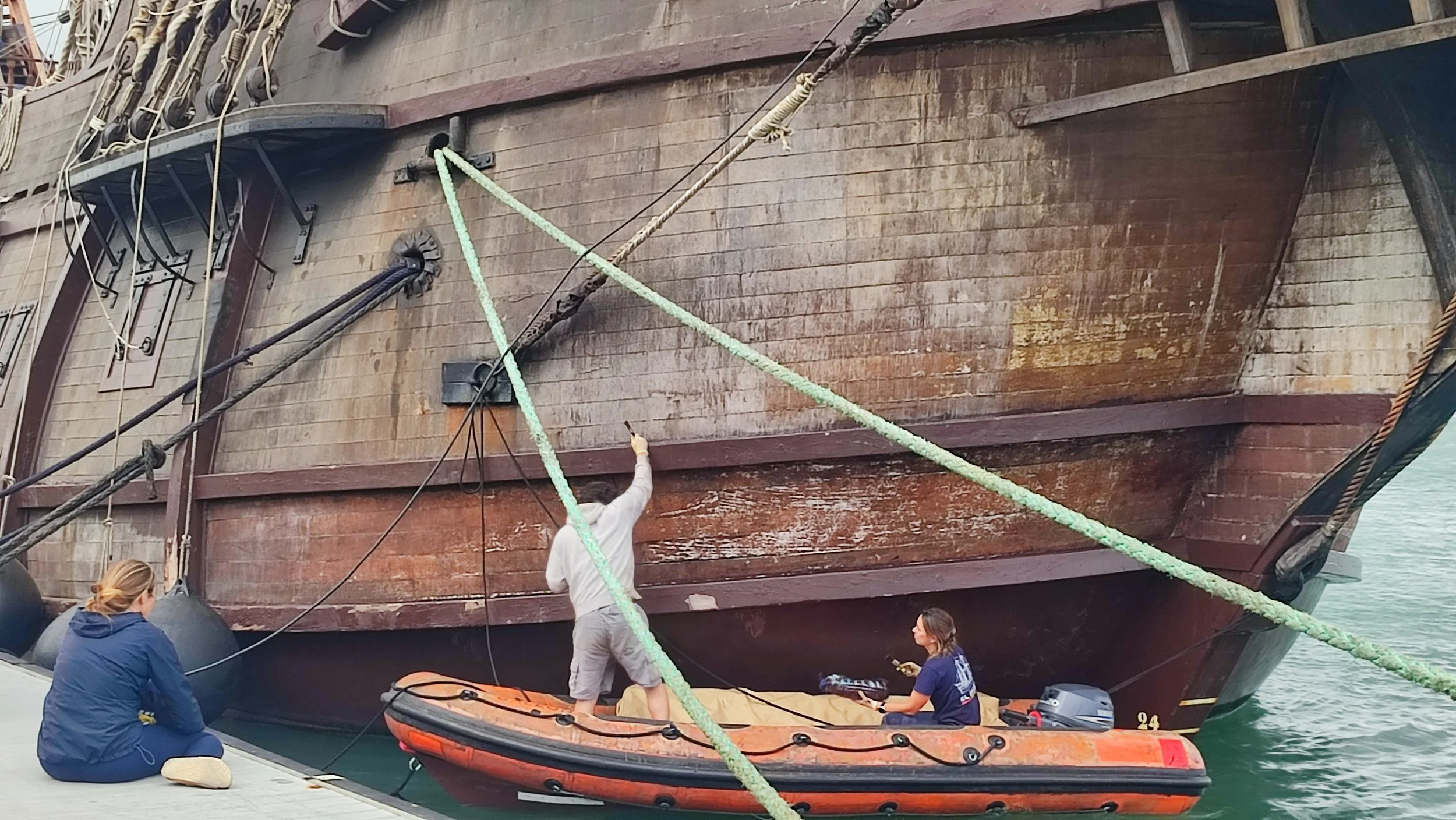 Two people on a dock near a large wooden ship. One person is sitting on the dock, and the other is in a small orange boat, pointing towards the ship's hull.