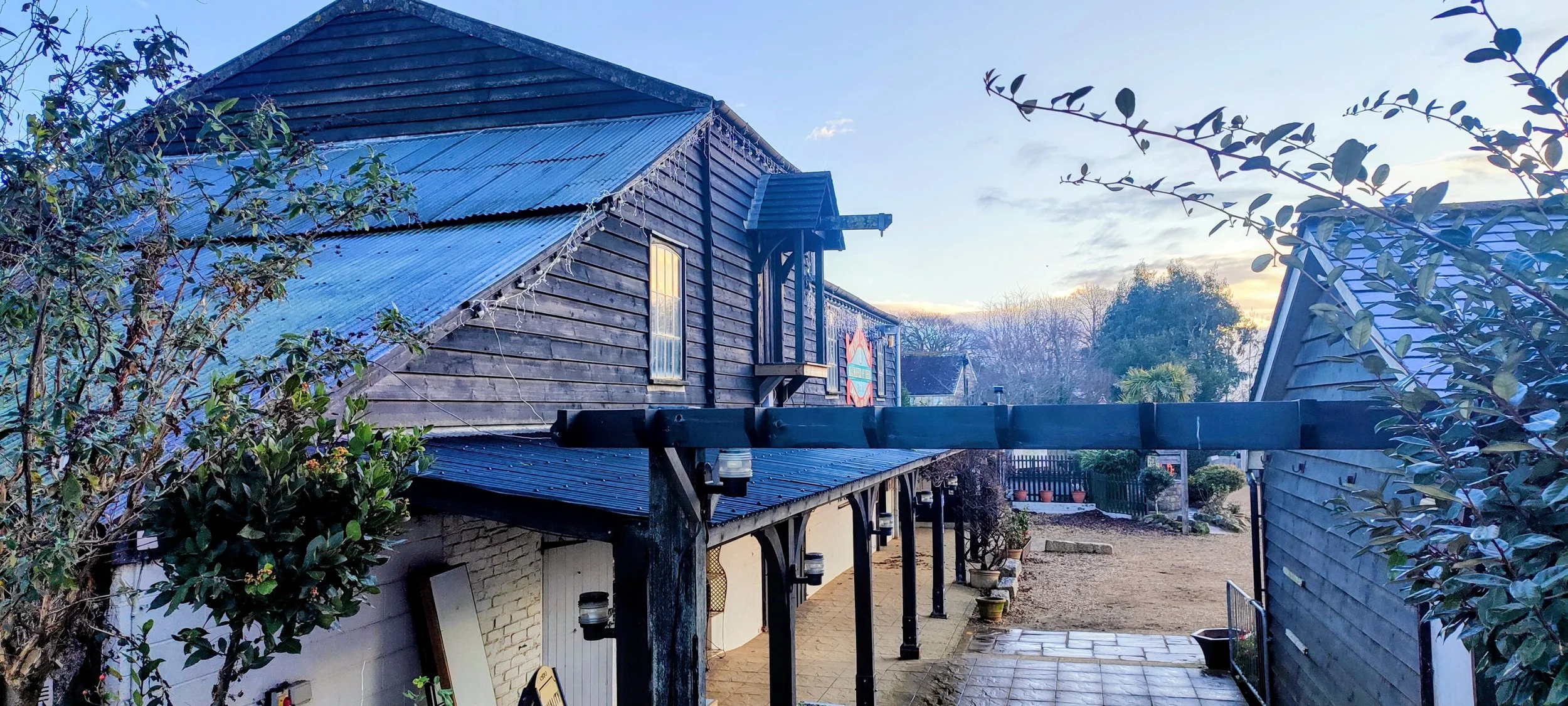 A rustic two-story building with weathered wooden siding and a corrugated metal roof, surrounded by patio with potted plants, trees, and a fence, under a blue sky with clouds.