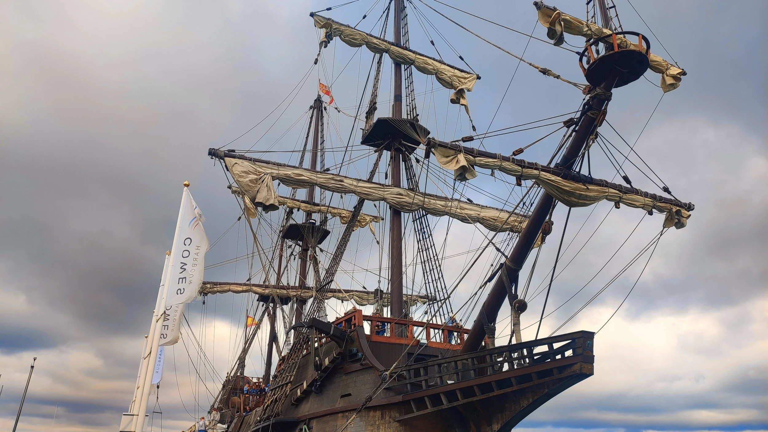 Replica of a historic sailing ship with multiple masts and furled sails, docked at a harbor under a cloudy sky.