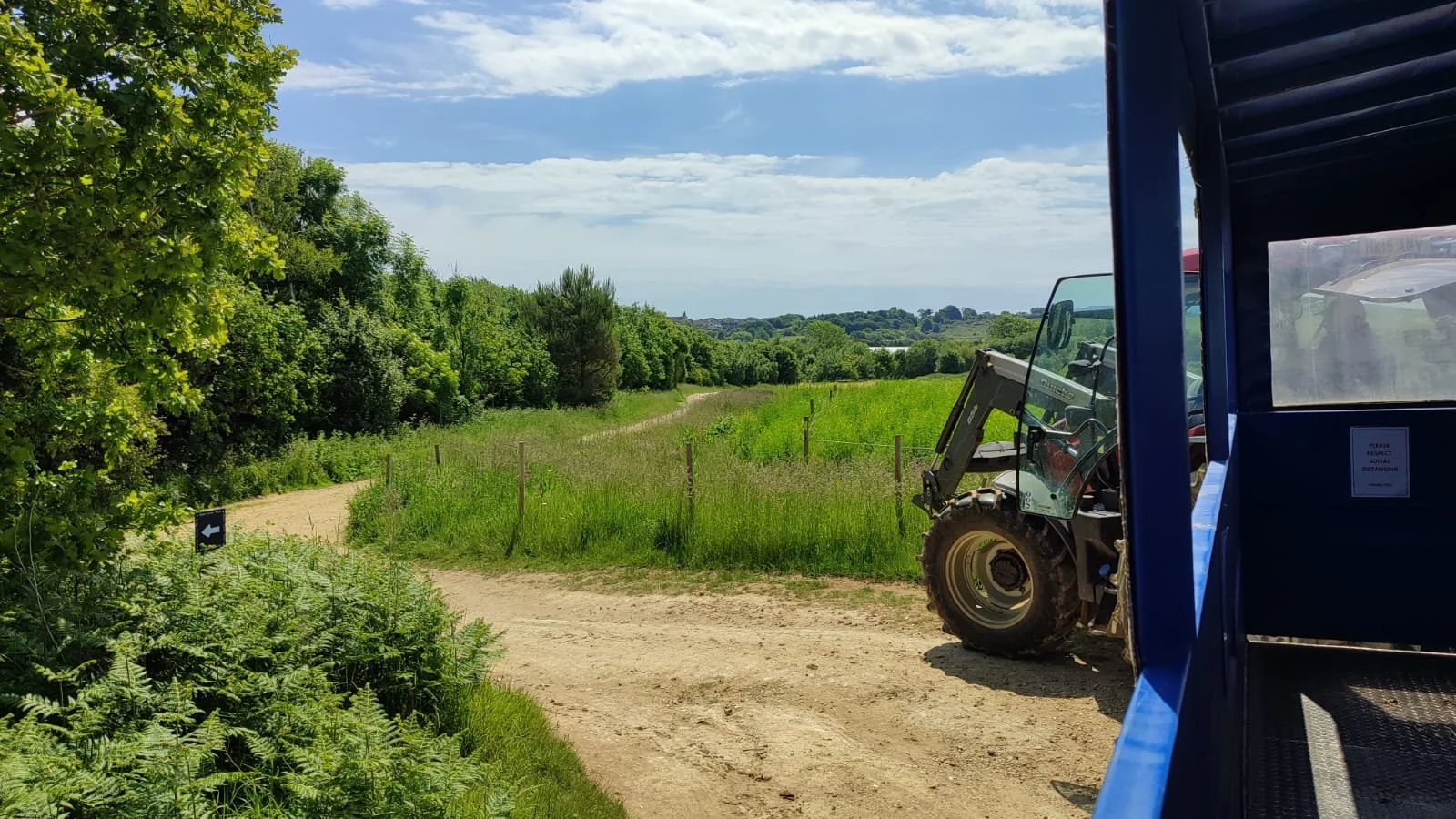View of a dirt trail in a green, rural area with trees and grass. A small construction vehicle is partially visible on the right side, parked next to a blue structure. The sky is partly cloudy with some scattered clouds.