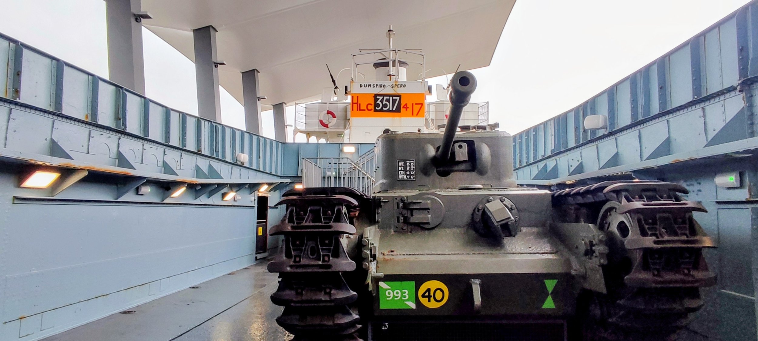 A military tank on a ship's deck with a large gun barrel pointing forward, surrounded by blue railings and a white superstructure in the background.