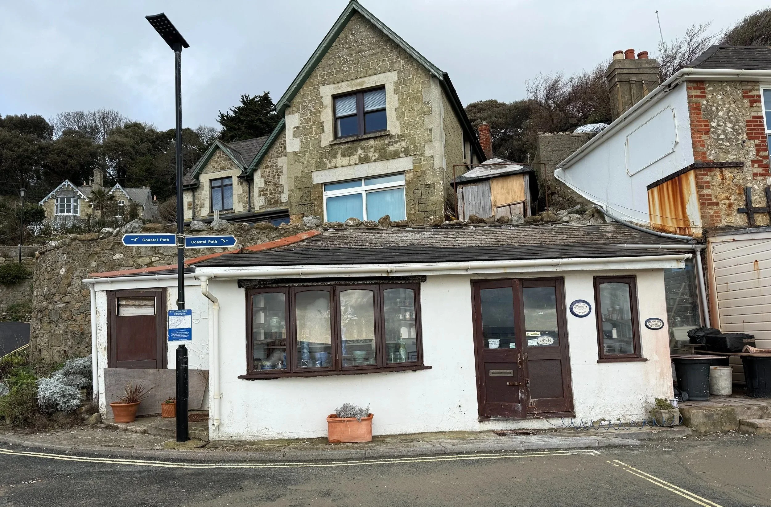 A small white building with brown window frames and a door, situated on a street corner, with potted plants outside. Above, there are houses on a hill made of stone and brick, with a cloudy sky overhead.