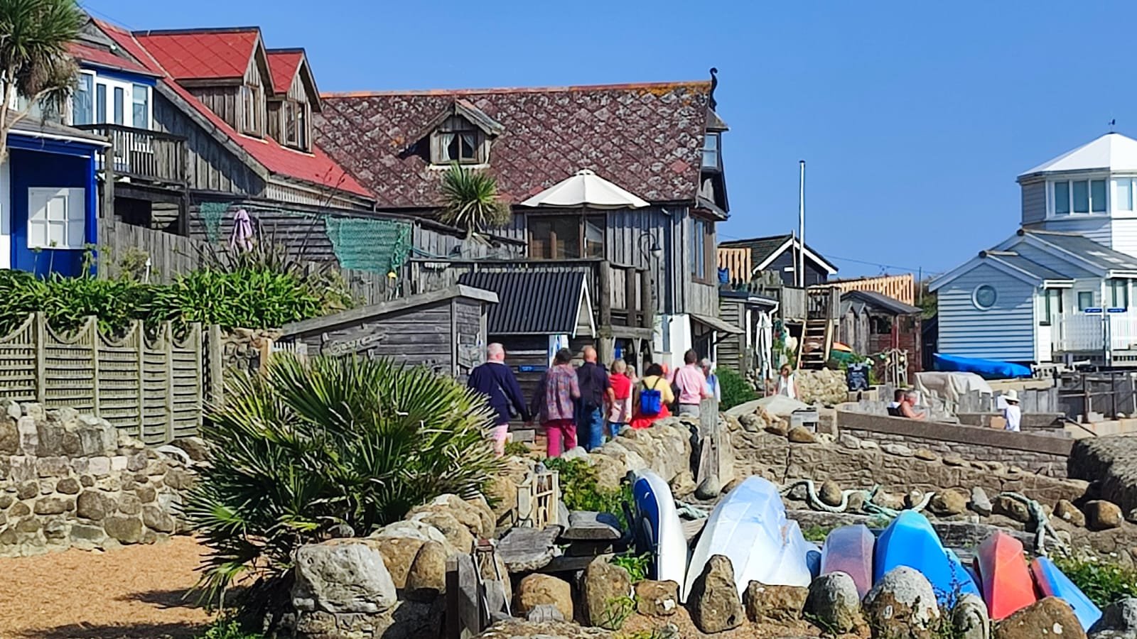 A group of people walking along a pathway near houses and boats by the water on a sunny day.