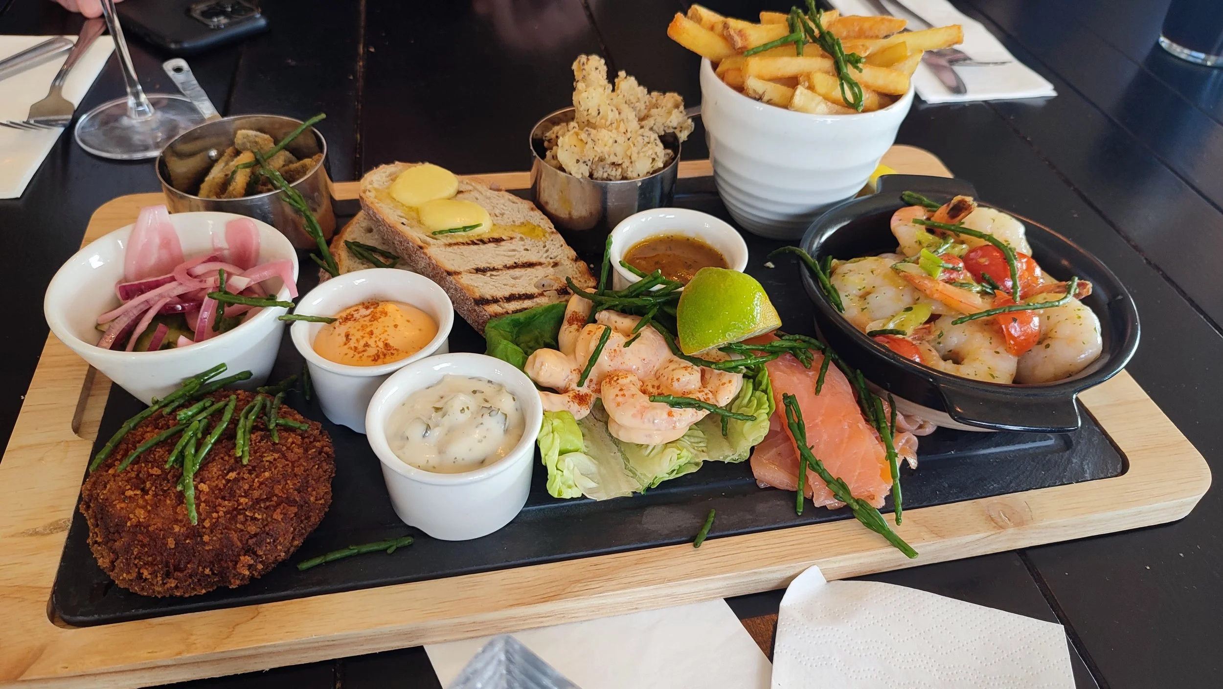 A seafood platter with grilled shrimp, smoked salmon, and cooked scallops served with various dipping sauces, grilled bread, and sides of French fries, fried calamari, pickled onions, and vegetable dishes on a wooden tray.
