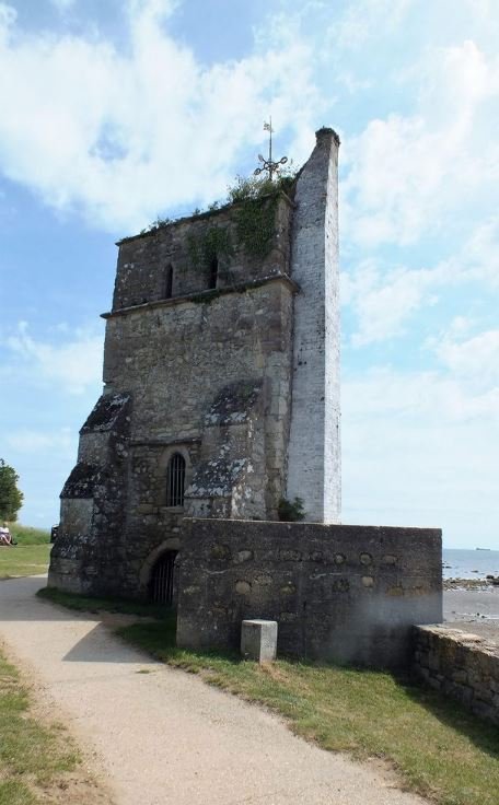 Old stone tower with small windows, located by a body of water with a stone wall in front and a dirt path leading up to it.