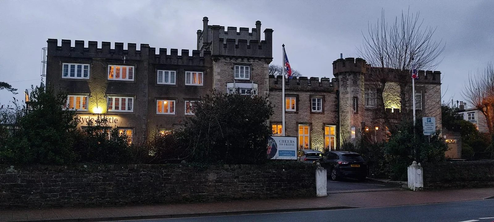 A castle-like building with illuminated windows, two flags on poles, and a parking lot in front, set against a cloudy evening sky.