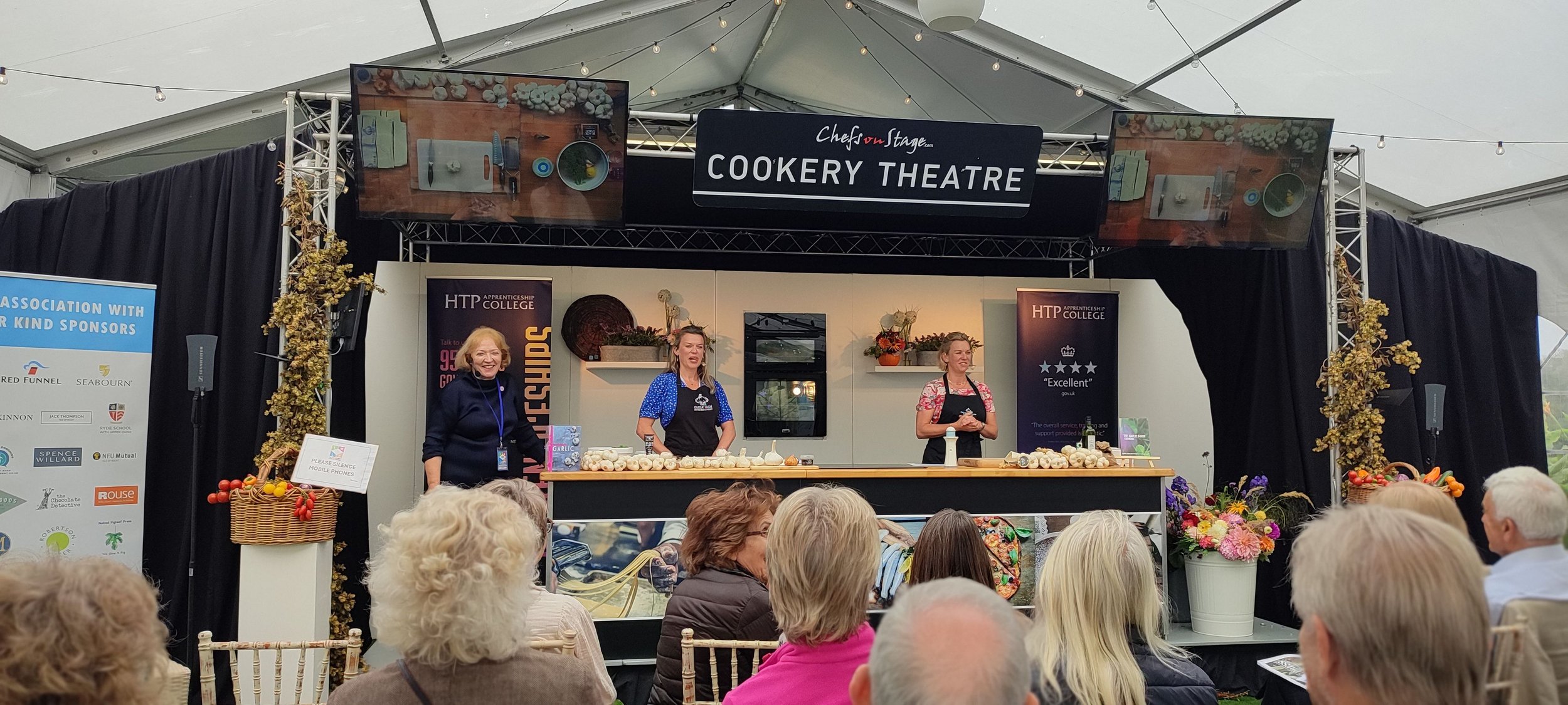 Three women on a cooking stage teaching a class, with an audience seated in front, and two screens displaying food images overhead.
