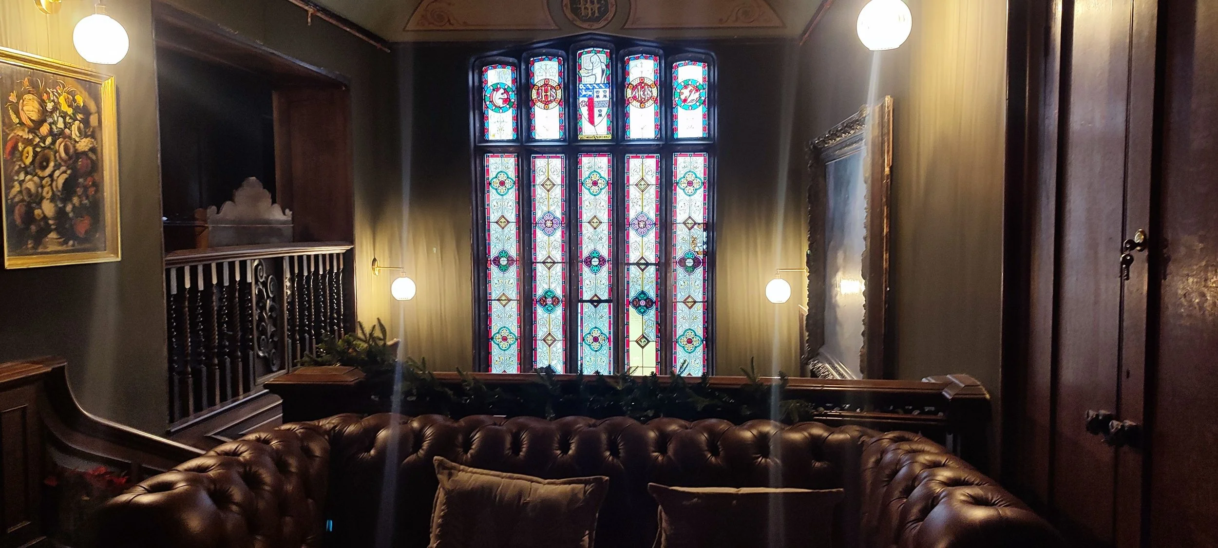 Interior of a room with a large stained glass window, a leather sofa, framed paintings, and soft lighting from wall-mounted lamps.