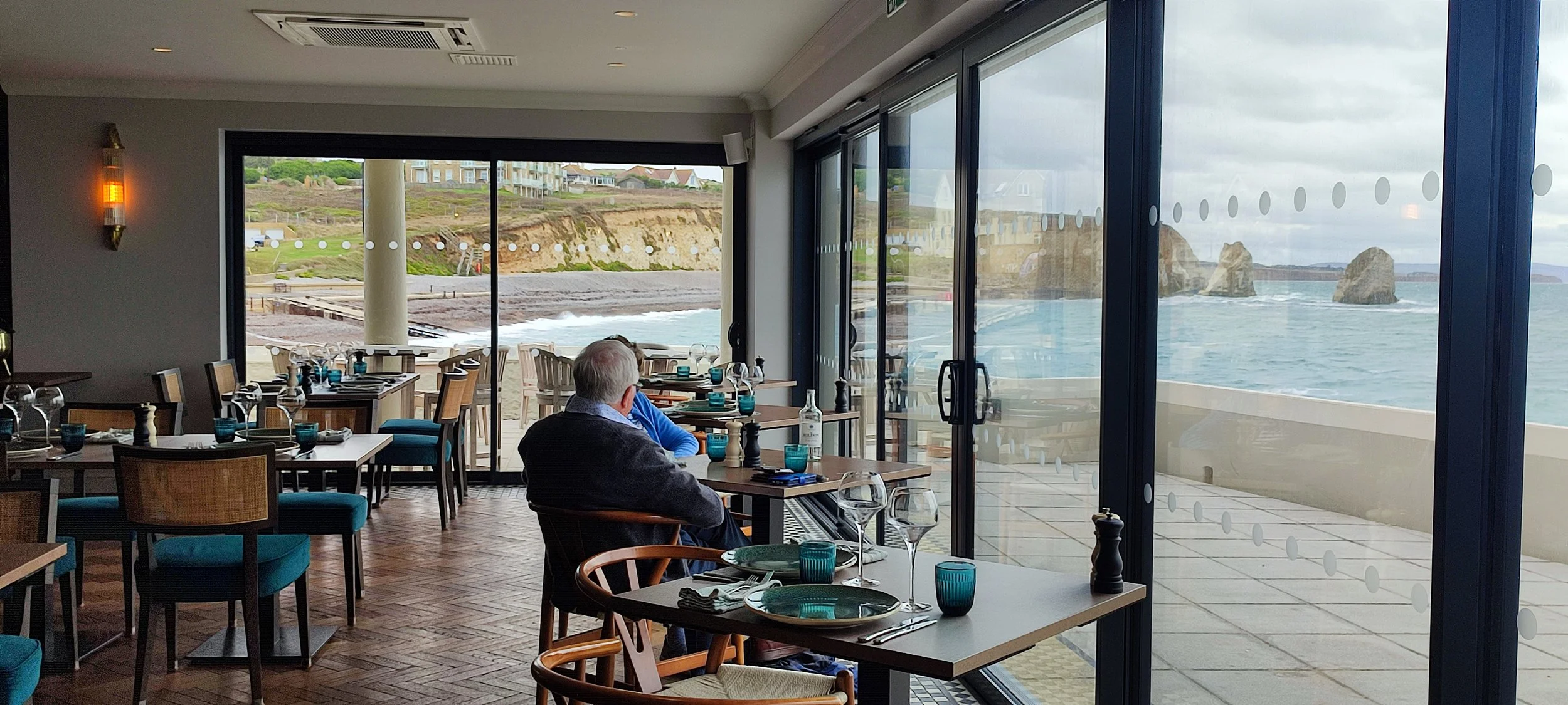 Indoor restaurant overlooking a beach and ocean with large glass windows and tables set with plates, glasses, and utensils. A few patrons are seated, enjoying the view of cliffs, rocks, and sea outside.