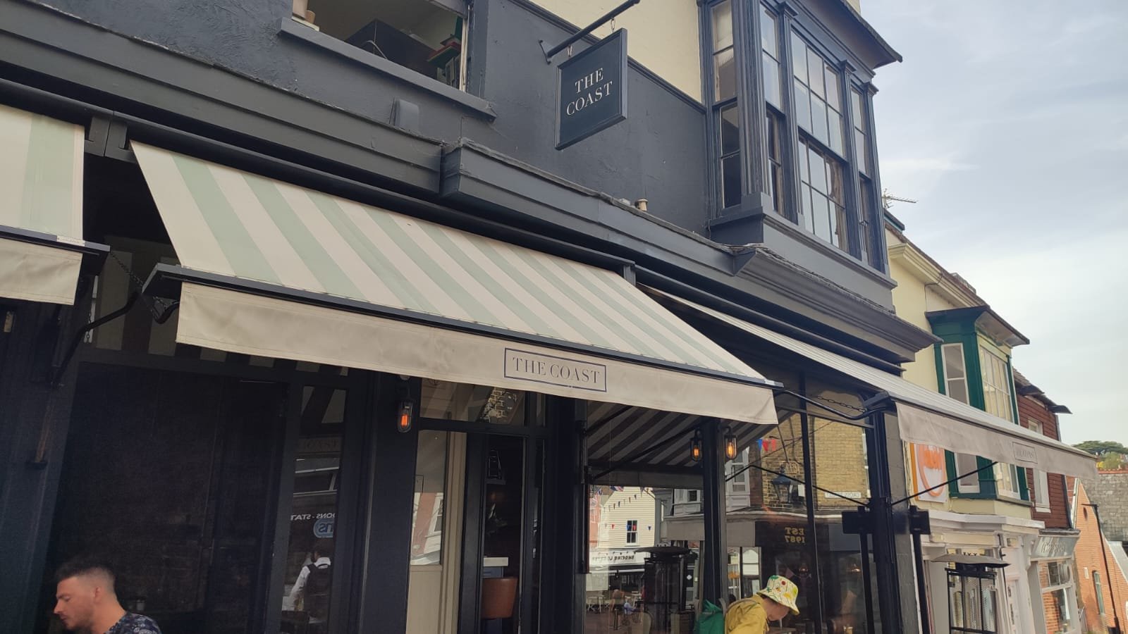 Exterior view of a restaurant named 'The Coast' with black framing, large windows, striped awnings, and an awning sign. There are people walking by, including a person in a yellow shirt with a colorful hat.