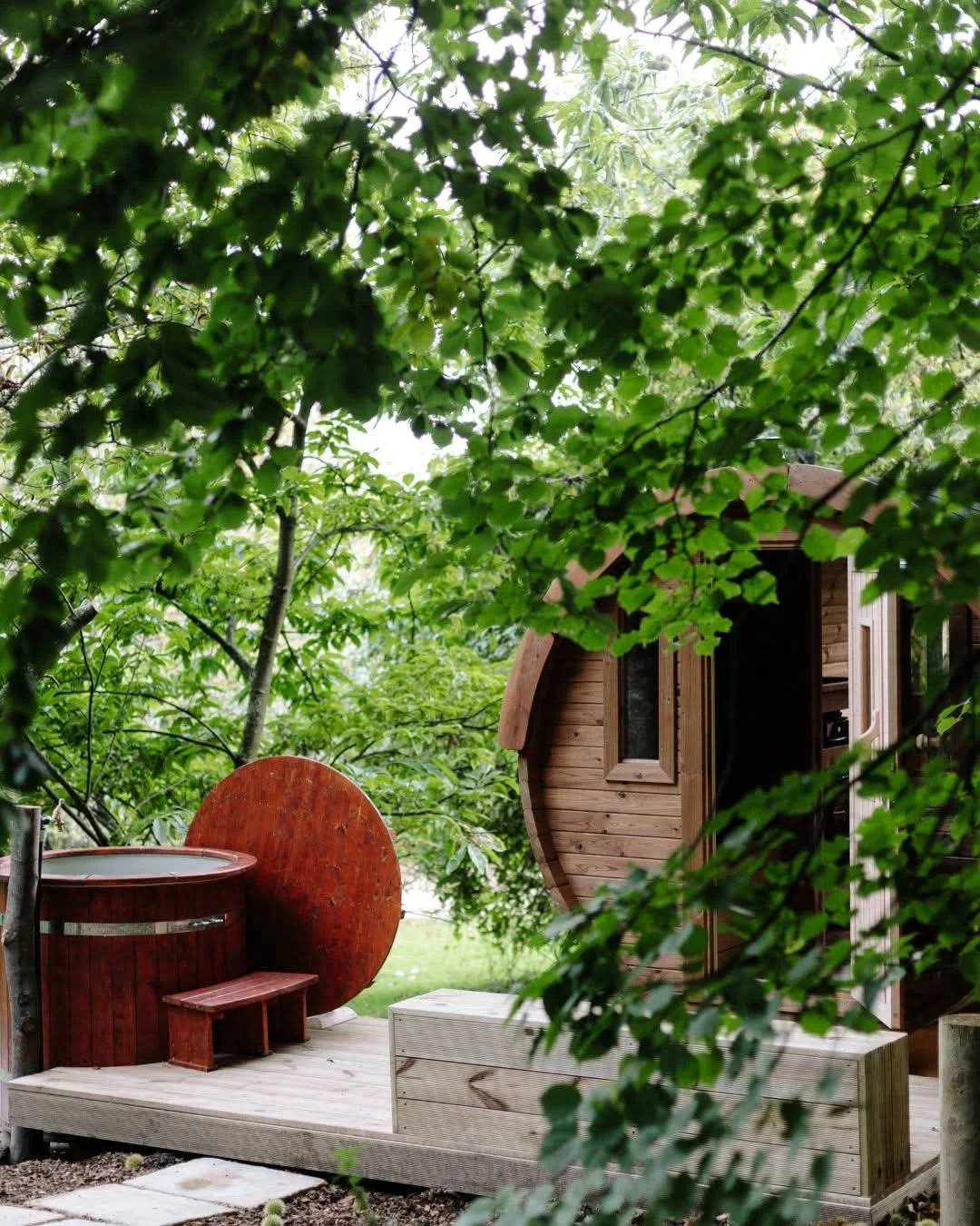 A wooden tiny house surrounded by lush green trees, with a small deck featuring a hot tub and a red wooden stool.