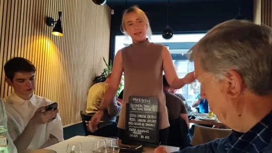 A young woman standing and speaking to a group at a restaurant table while others are sitting and listening.