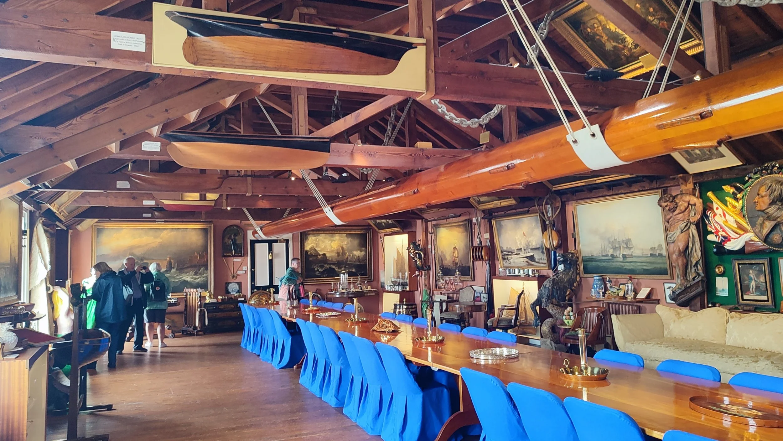 A room decorated with nautical-themed paintings and sculptures, with large wooden canoes hanging from the ceiling. There are several art pieces, a long wooden table with blue chairs, and a group of people viewing the art.