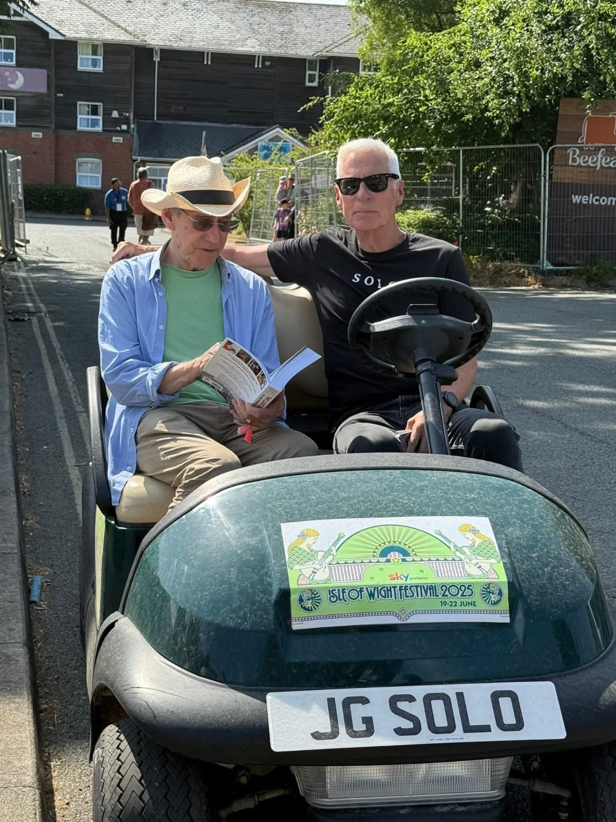 Two men sitting in a golf cart, one reading pamphlet and the other wearing sunglasses, with signs indicating the Isle of Wight Festival 2025, and a license plate reading JG SOLO.