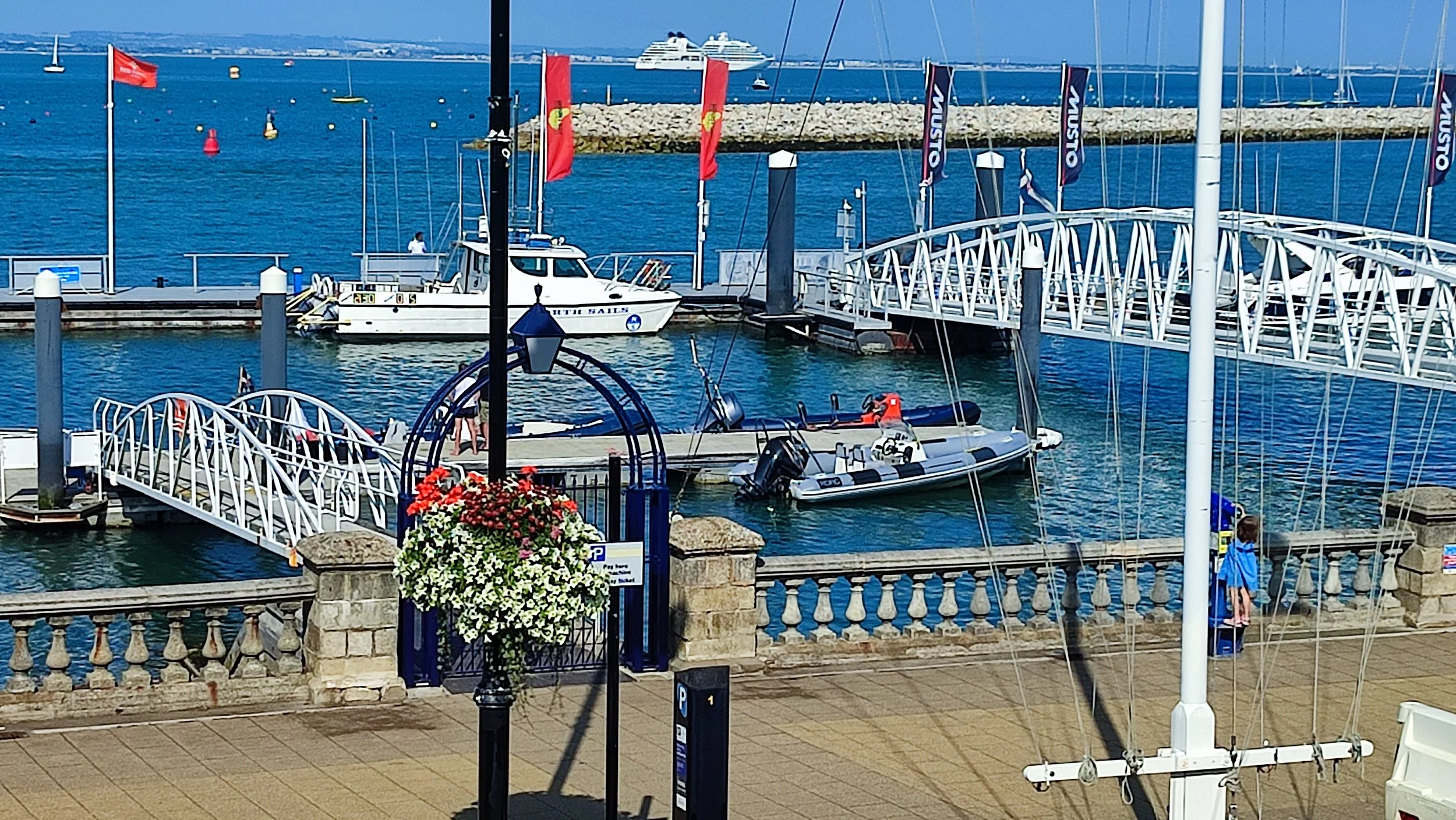 View of a marina with several boats docked, including a yacht and a speedboat. A stone and iron railing separates the marina from the paved walkway. There is a large cruise ship in the distance on the ocean, and a cruise ship on the harbor. Red and blue flags are displayed. A person with a blue jacket is standing near the railing.