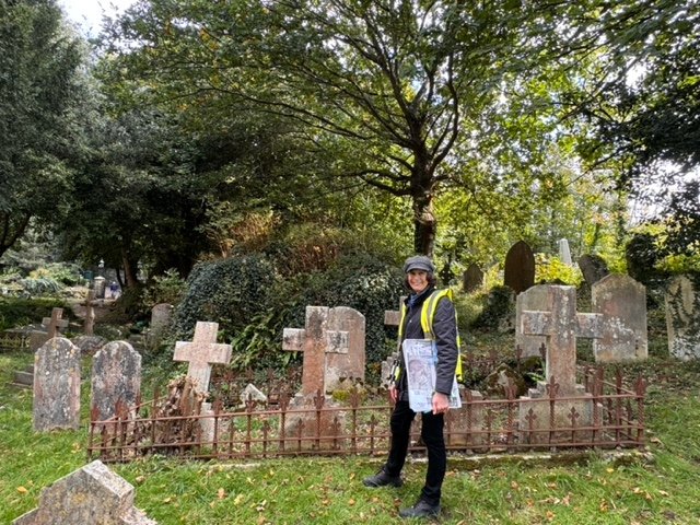 A person wearing a yellow safety vest and a helmet standing in front of old gravestones in a cemetery with trees and greenery in the background.