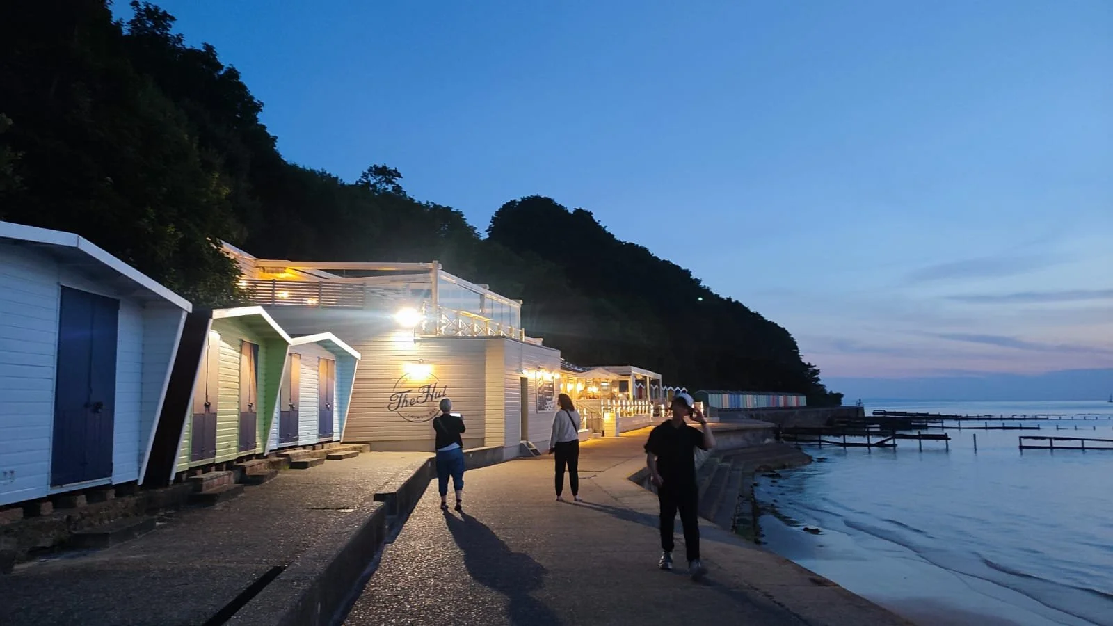 A seaside walkway at dusk with colorful beach huts on the left and a calm ocean on the right. Three people are walking along the path, some wearing hats and carrying bags. The background features a hillside covered with trees and a clear sky transitioning from day to night.