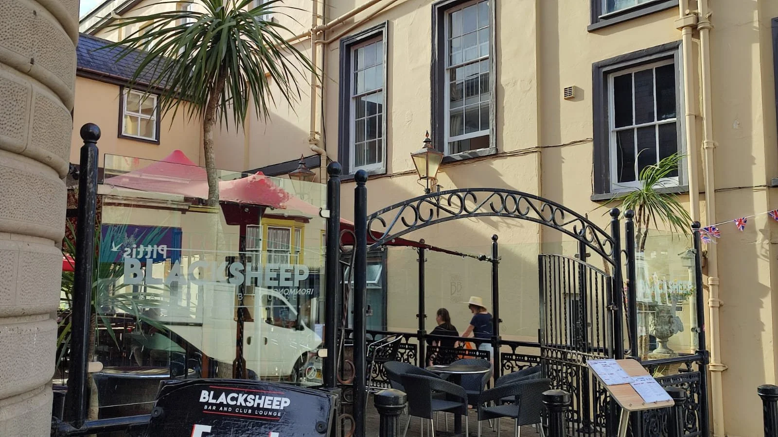 An outdoor patio area of a bar and lounge with black wrought iron fencing, black chairs, and tables. There are two women sitting and talking, one wearing a sun hat. A palm tree and glass panels with the name "Black Sheep" are visible.