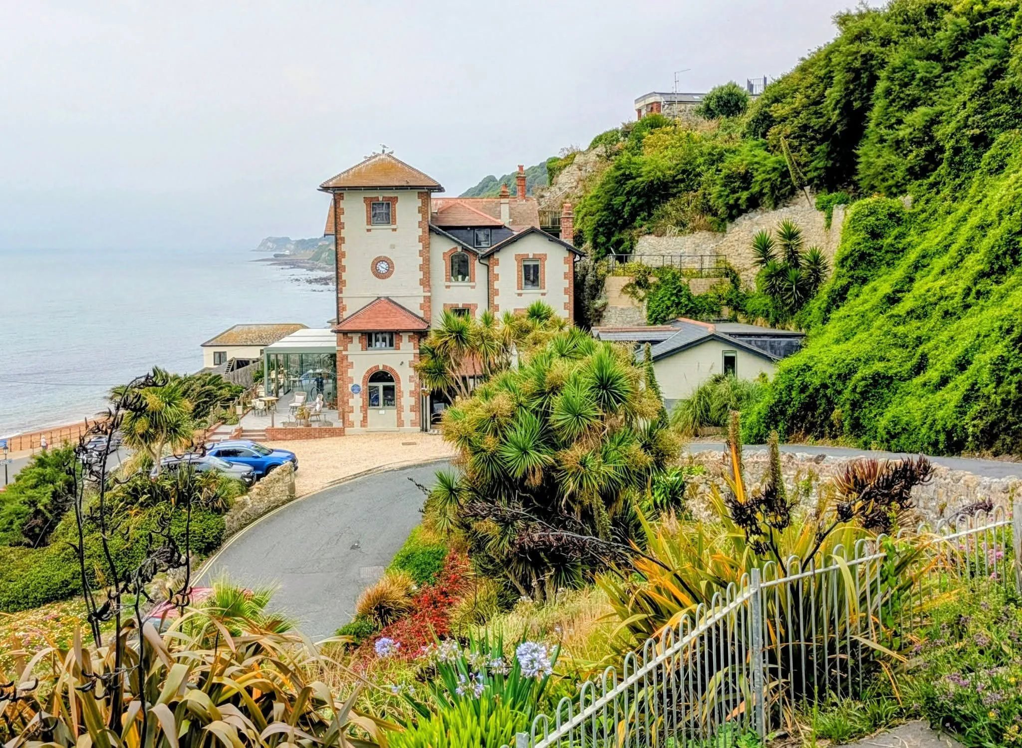 A large, multi-story house on a hill with a view of the ocean, surrounded by lush green plants and trees, with a winding driveway, parked cars, and a garden.