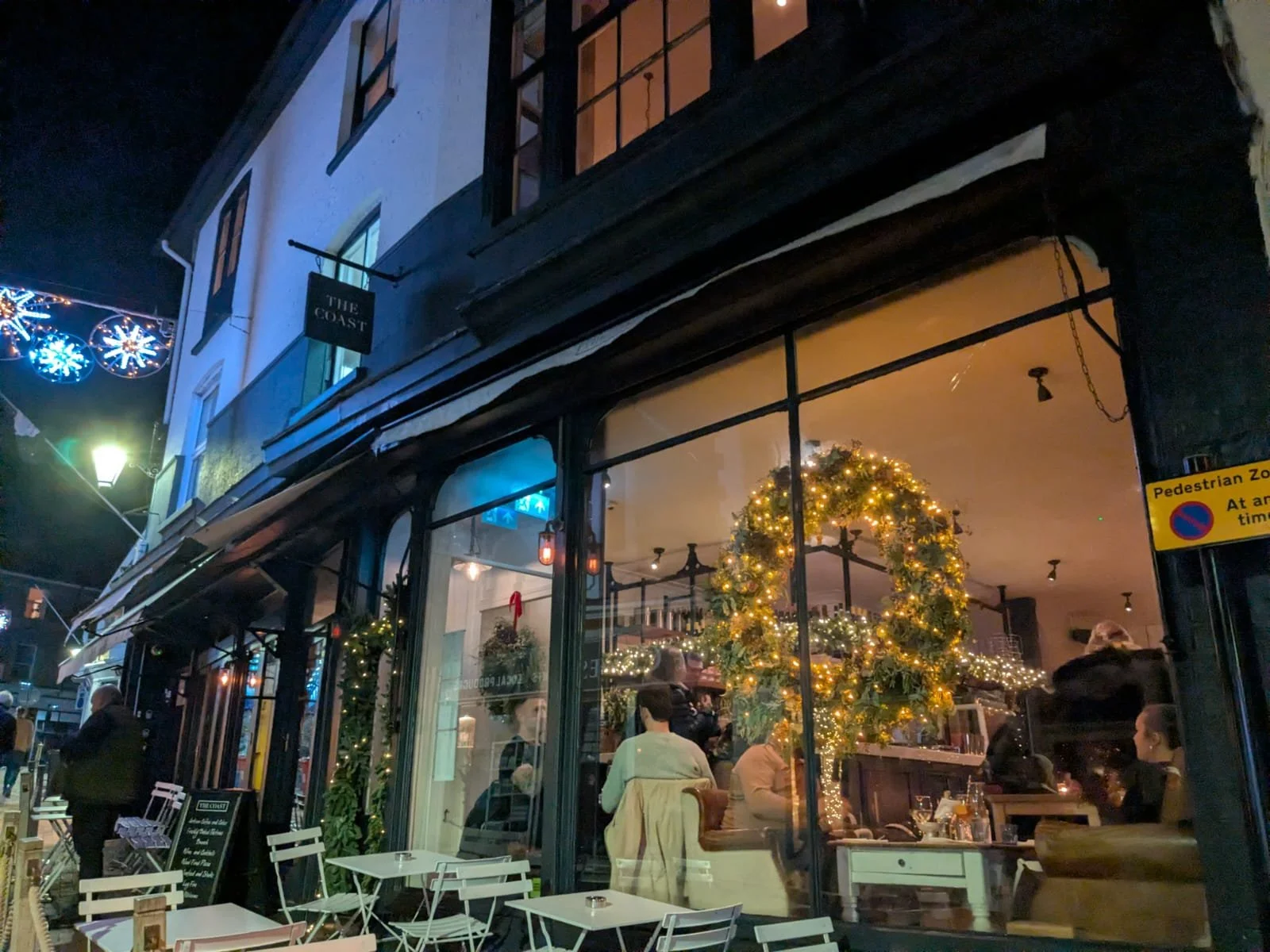 Nighttime view of a restaurant decorated with Christmas lights, wreaths, and a large Christmas tree inside. People are sitting and dining inside, visible through large glass windows, with some outdoor seating available. The scene is festive with holiday decorations and warm lighting.