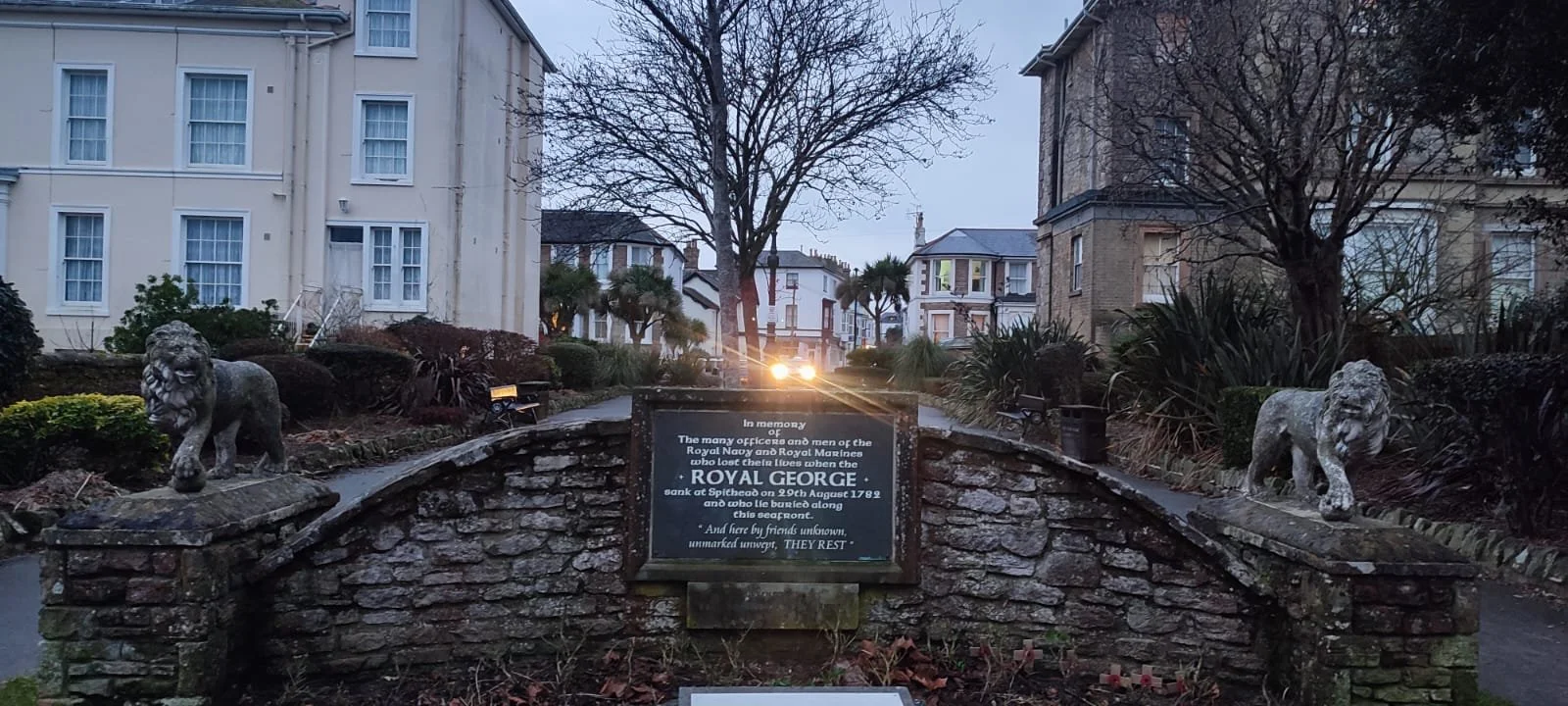 Memorial plaque on a small brick bridge with stone lion statues on each end, situated in a garden with leafless trees and surrounding buildings at dusk.