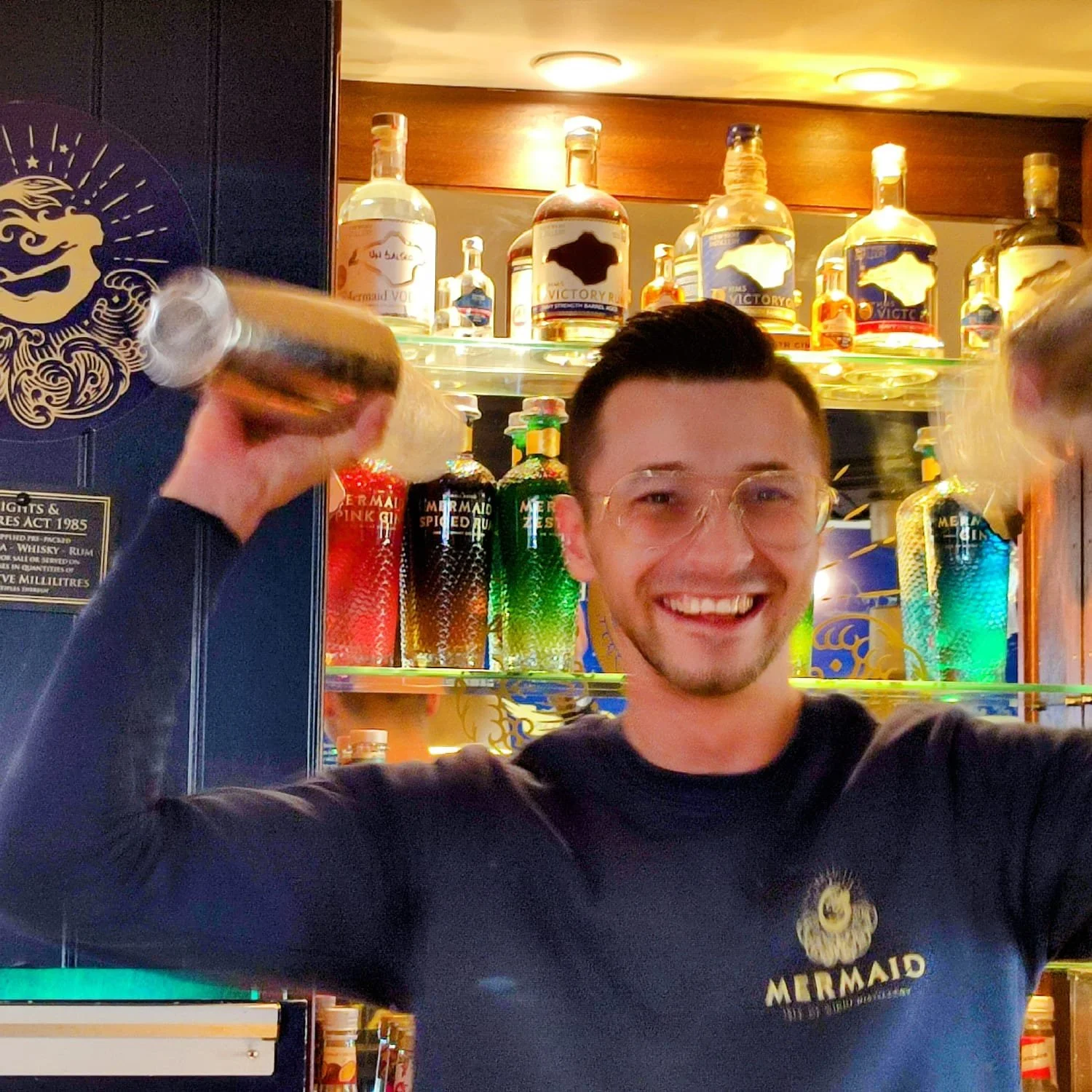 A smiling young man with short dark hair and glasses, wearing a Mermaid-themed shirt, holding a bottle in each hand, inside a bar with colorful bottles of alcohol on shelves behind him.