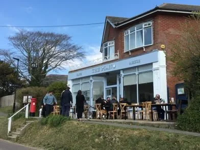 People sitting and standing outside a cafe with outdoor seating, a red mailbox, and a brick building with large front windows.