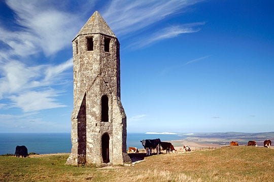 A stone tower standing on grassy land overlooking the ocean with a blue sky and clouds.