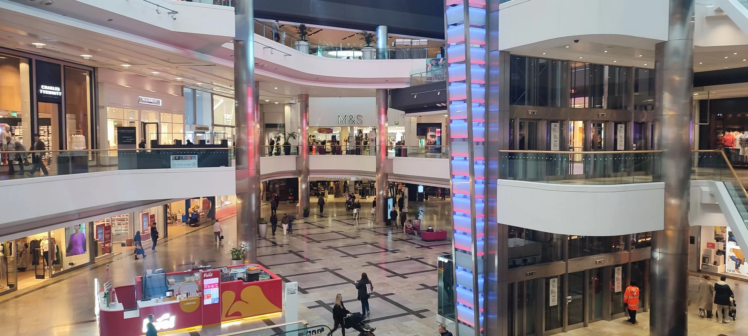 Interior of a multi-level shopping mall with various retail stores, a central open area, an elevator with blue lighting, and shoppers walking around.
