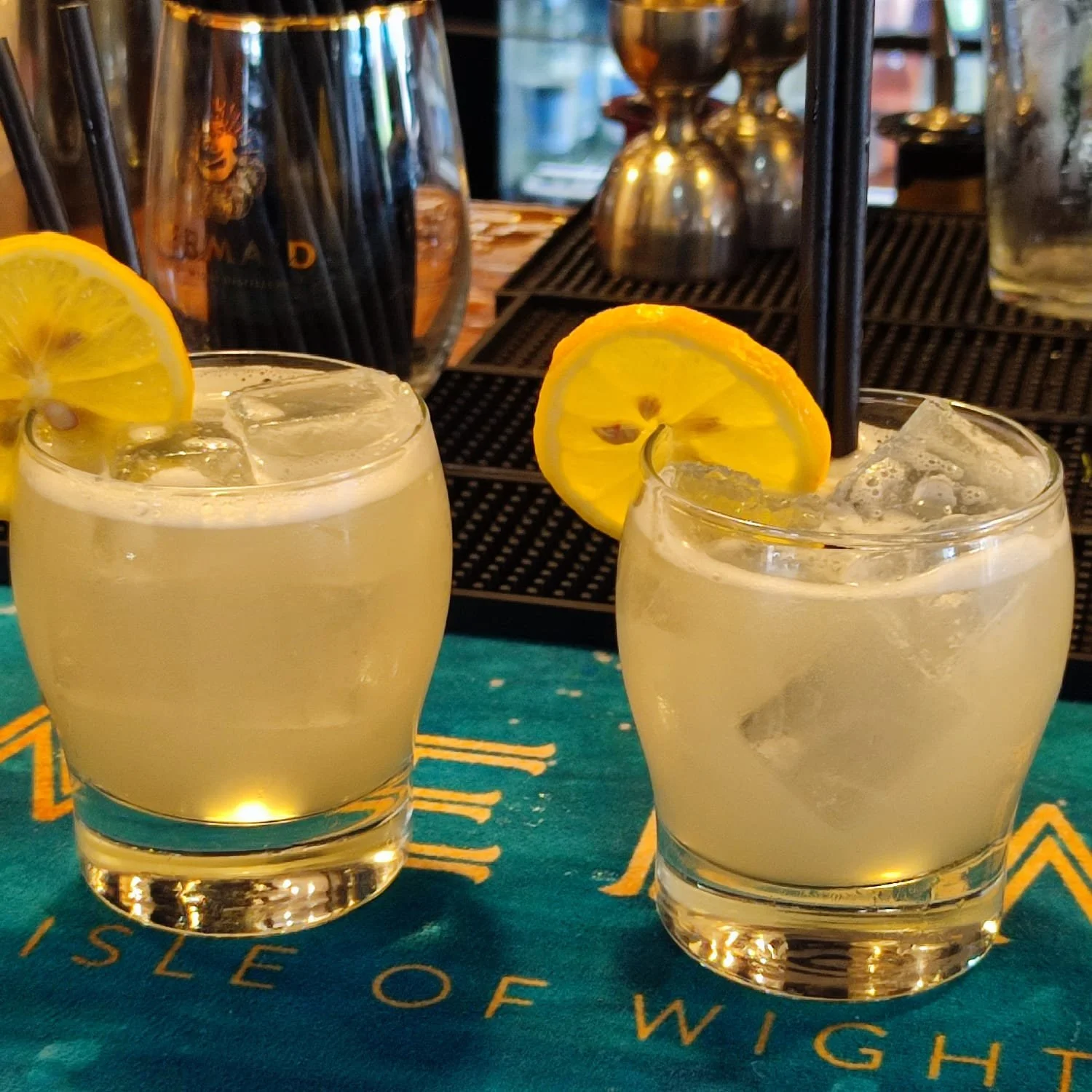 Two glasses of lemonade with lemon slices and ice on a blue cloth with yellow writing, in front of bar equipment.