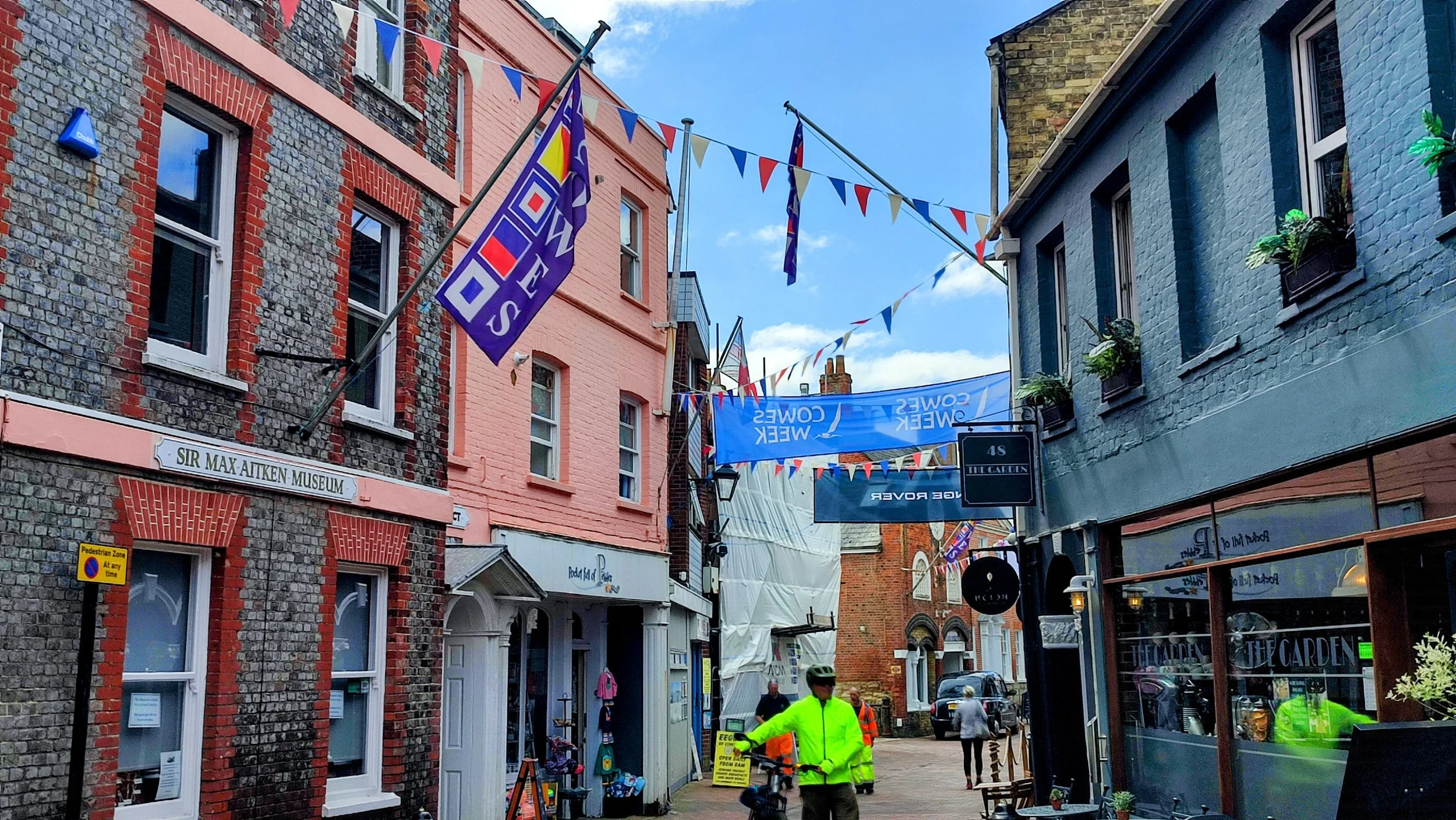 A narrow street in an urban area decorated with colorful flags and banners, with buildings on both sides including the Sir Max Aitken Museum on the left and a shop with large windows on the right; people are walking and a cyclist in a neon yellow jacket is in the foreground.