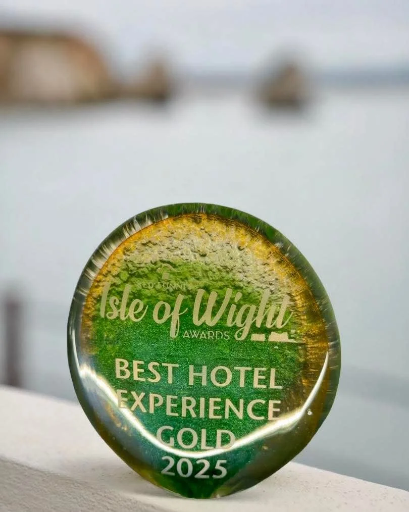 Glass award with the words "Isle of Wight Awards 2025 Best Hotel Experience Gold" etched on it, placed outdoors near a body of water with blurred boats in the background.