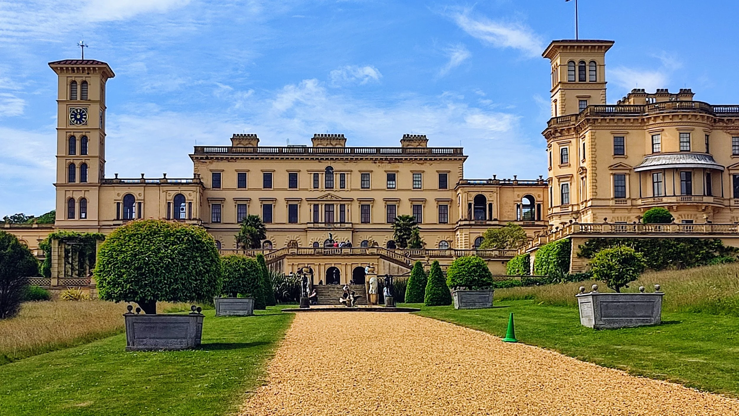 A grand historic castle with beige walls, towers, and columns, surrounded by a well-manicured lawn with trimmed bushes, potted plants, a gravel walkway leading to the entrance, and a bright blue sky overhead.