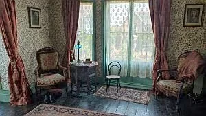 Living room with floral curtains, a window with lace curtains, two upholstered chairs, a small table with decorative items, and a patterned rug on the wooden floor.