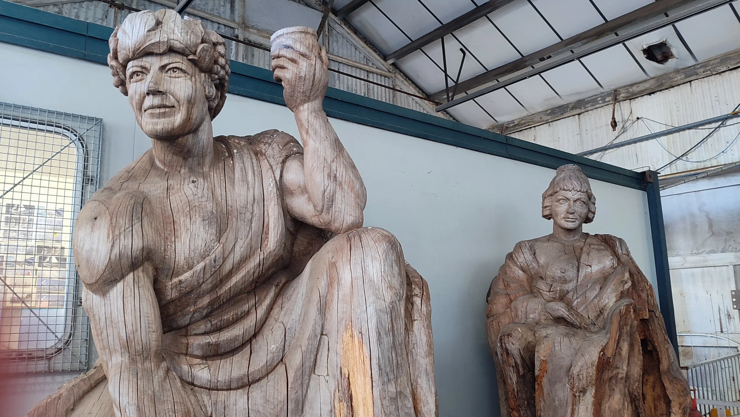 Two large wooden sculptures of women with detailed facial features and hairstyles, made from carved tree trunks, inside a workshop or storage area.