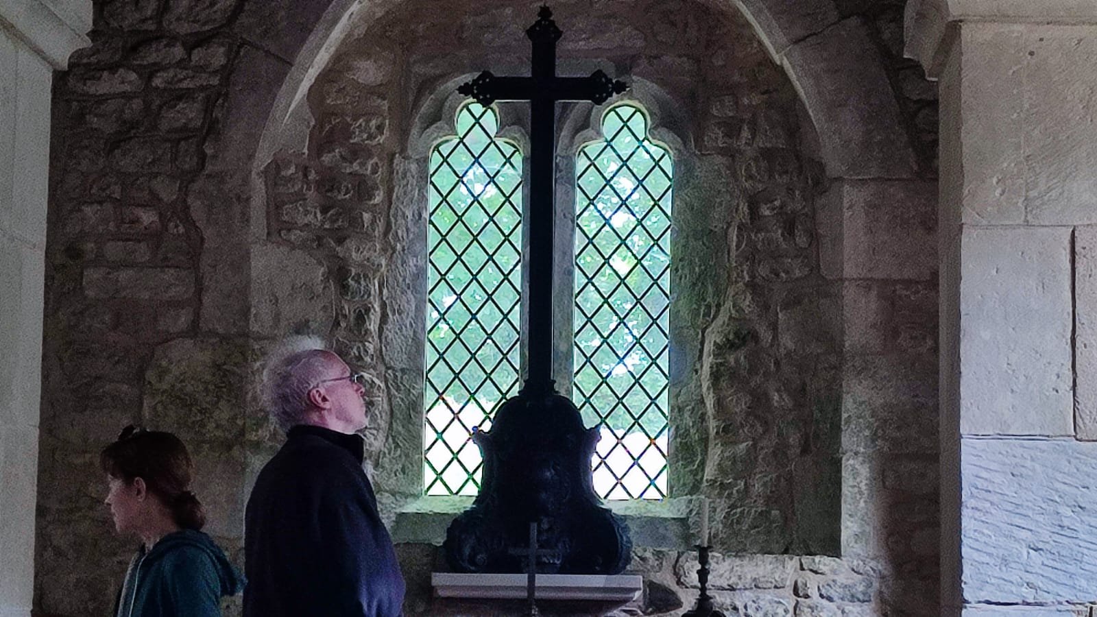 People inside a stone building with a large window covered by a decorative iron grille. A man with glasses and a woman are looking at a black altar with a crucifix at the center.