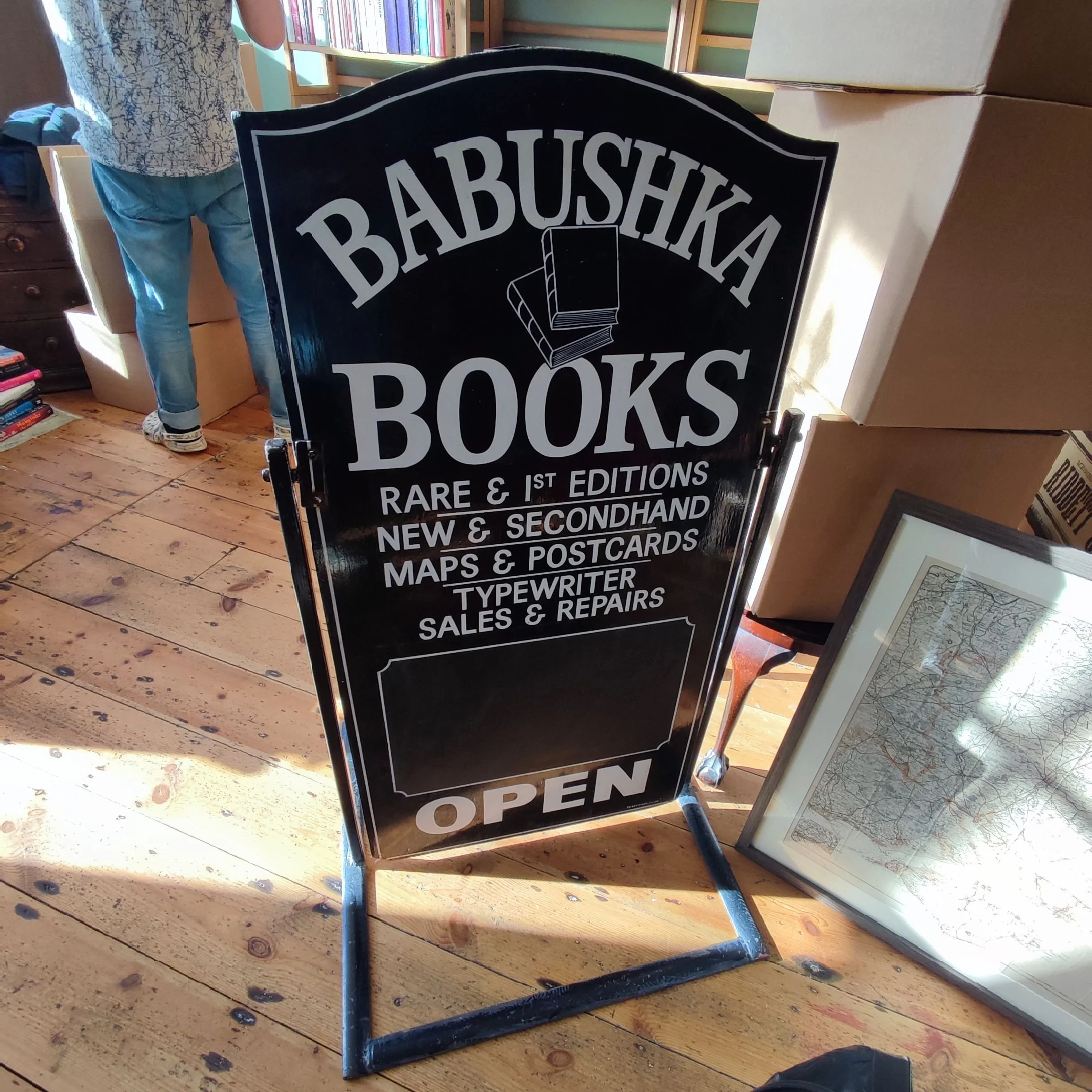 A black standing signboard with white text advertising a bookstore called 'Babushka Books'. The sign indicates they sell rare and first editions, new and secondhand books, maps and postcards, and offer typewriter sales and repairs. The sign also states it is open. A framed map is leaning against boxes on the right, and a person is partially visible on the left, standing near bookshelves.