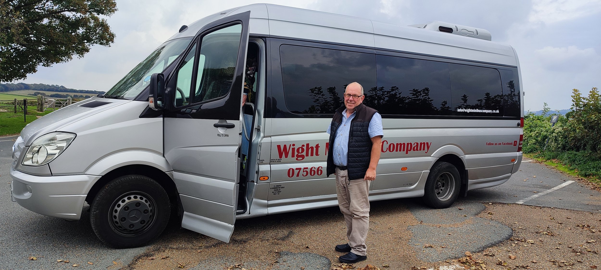 A man standing in front of a silver minibus with the text 'Wight Minibus Company' and a contact number on the side. The man is smiling, wearing glasses, a light blue shirt, and a black vest. The minibus is parked on a paved area with some leaves on the ground, and there are trees and fields in the background under a cloudy sky.