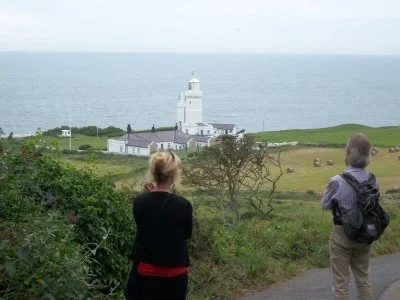 Two people standing on a hillside overlooking a coastline with a white lighthouse and buildings, under an overcast sky.