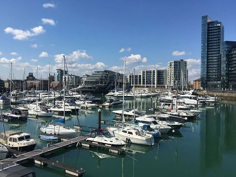 Marina with numerous docked sailboats and yachts against a backdrop of modern buildings and a blue sky with scattered clouds.