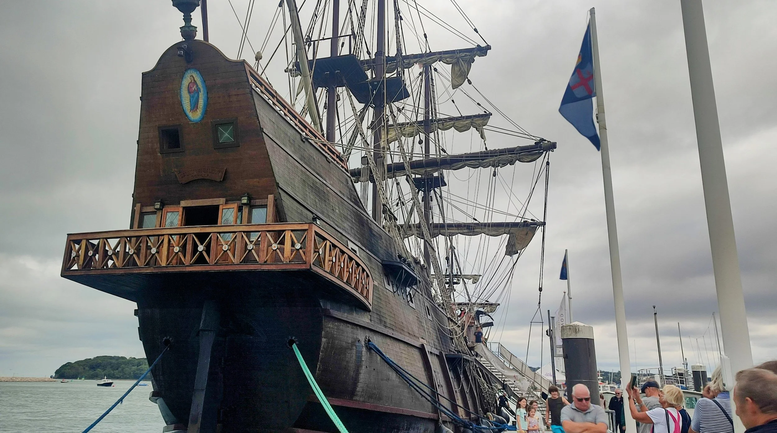 A large, historic wooden pirate ship docked at a marina with people walking around and taking photos; cloudy sky.