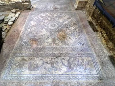 Ancient mosaic floor with geometric and animal patterns, partially restored and surrounded by stone walls.