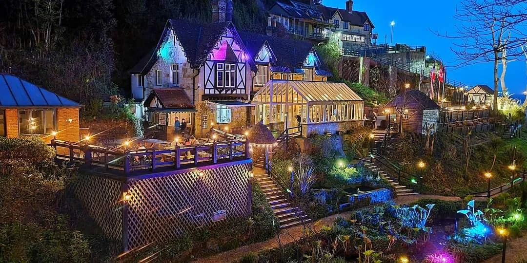 Night view of a hillside property with multiple illuminated buildings and gardens, including a house with a greenhouse and outdoor dining area.