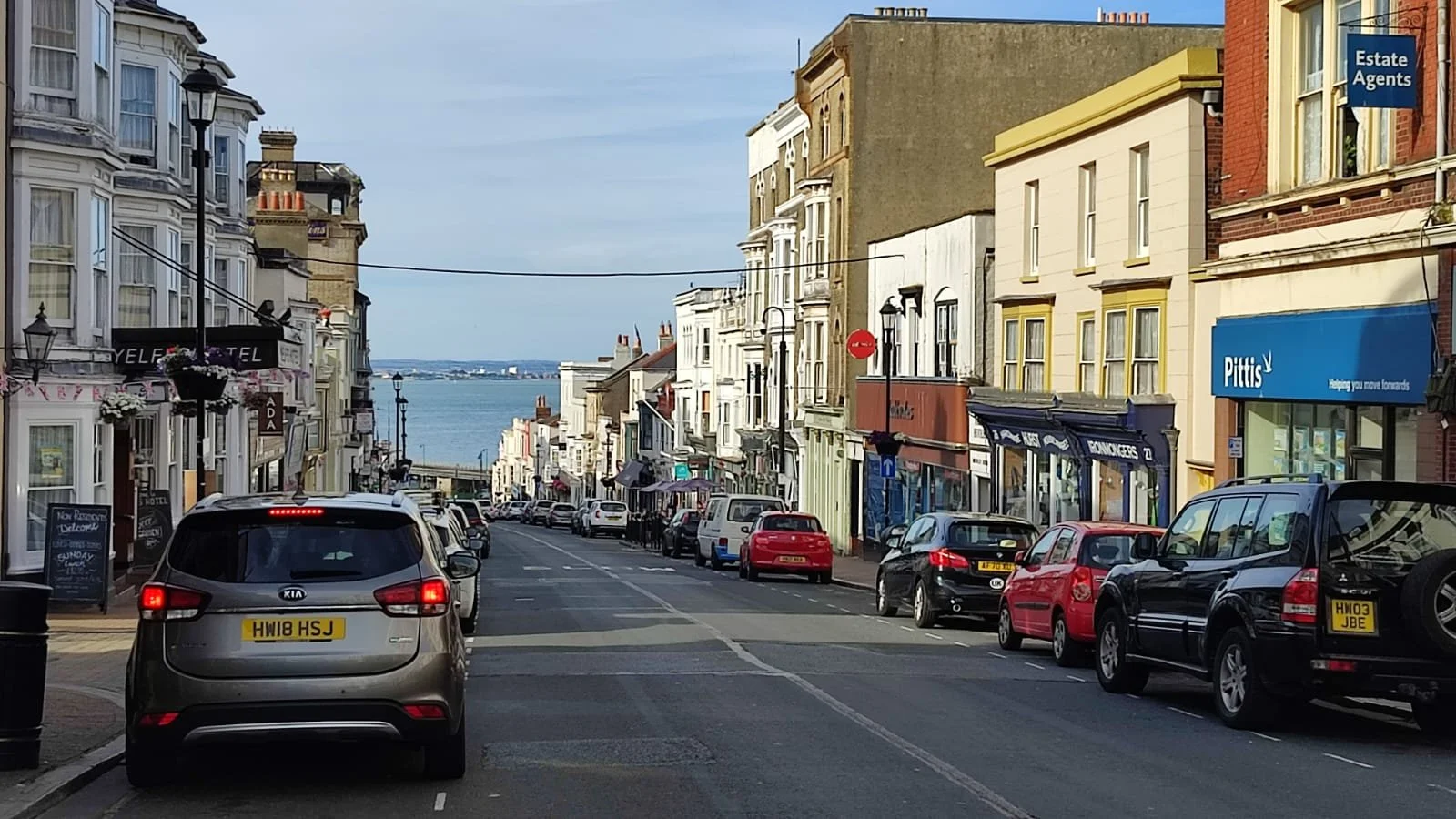 Street scene with parked cars, storefronts on both sides, with a view of water and distant land in the background.