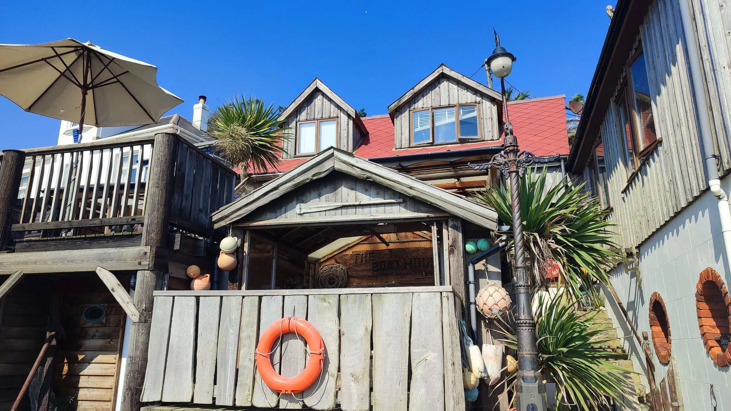 Wooden building with multiple levels and a red roof, outdoor deck with a large beige umbrella, potted plants, and a vintage lamppost, against a clear blue sky.