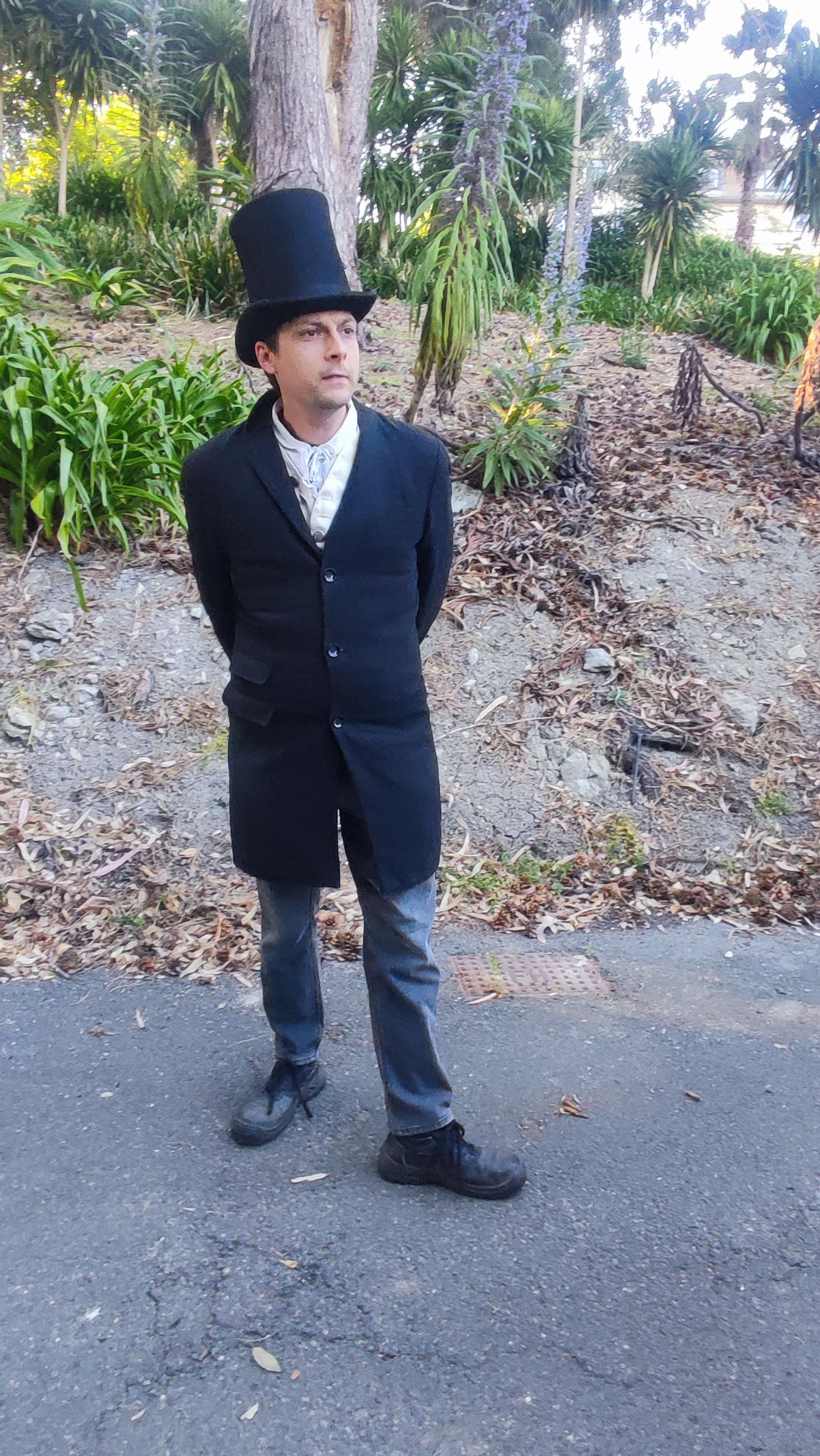 A person dressed in vintage 19th-century attire, wearing a tall black top hat and a long black coat, standing outdoors on a paved area with trees and plants in the background.